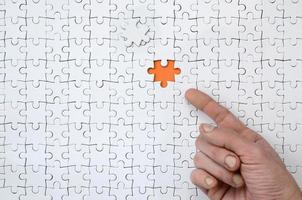 The texture of a white jigsaw puzzle in the assembled state with one missing element, forming an orange space, pointed to by the finger of the male hand photo