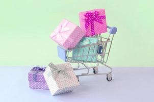 Small gift boxes of different colors with ribbons in shopping cart on a violet and blue pastel background photo