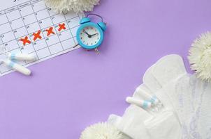 Menstrual pads and tampons on menstruation period calendar with blue alarm clock and white flowers photo