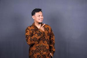 Portrait of a happy young man in batik indonesia pointing fingers away at copy space isolated over gray background photo