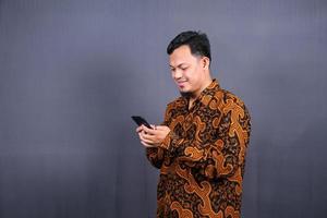 Portrait of asian young man wearing batik using cellphone on gray background photo