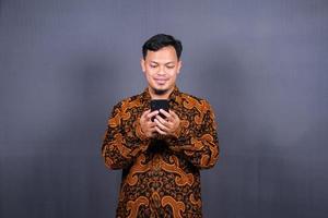 Portrait of asian young man wearing batik using cellphone on gray background photo