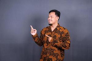 retrato de un joven feliz en batik indonesia señalando con el dedo el espacio de copia aislado sobre fondo gris foto