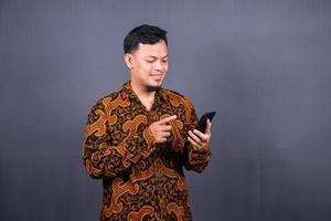 Portrait of asian young man wearing batik using cellphone on gray background photo