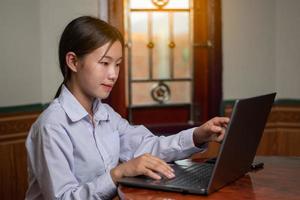 A student girl with white shirt student suit is touch the laptop in the house, using notebook and wireless internet on the desk. education and studying concept, sunrise light through the windows photo
