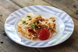 ensalada de papaya tailandesa en un plato, ensalada de papaya con caca, limón, pimienta, tomate rojo en un tazón blanco sobre una mesa de madera foto