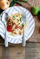 Thai papaya salad on a plate, papaya salad with poop, lemon, pepper, red tomato in white bowl on wooden table photo