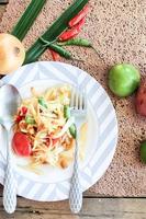 ensalada de papaya tailandesa en un plato, ensalada de papaya con caca, limón, pimienta, tomate rojo en un tazón blanco sobre una mesa de madera foto