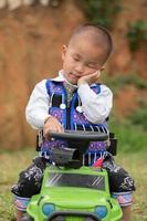 A kid sit on his green toy car, pose like as sleep, wear tradition clothe photo