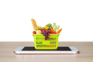 Fresh food and vegetables in green shopping basket on mobile smartphone on wood table top isolated on white background grocery online concept photo