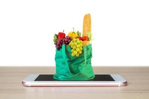 alimentos frescos y verduras en una bolsa de compras verde en un teléfono inteligente móvil en una mesa de madera aislada en un concepto en línea de comestibles de fondo blanco foto