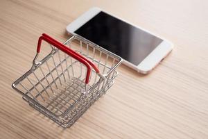teléfono móvil con cesta en el fondo de la mesa de madera concepto de compras en línea foto