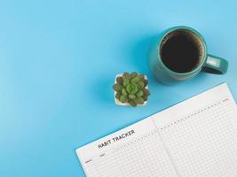 flat lay of habit tracker book with  blue  cup of black coffee and succulent plant pot on blue background with copy space. photo