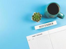 plano de libro de seguimiento de hábitos con calendario de madera noviembre, taza azul de café negro y maceta suculenta sobre fondo azul con espacio para copiar. foto