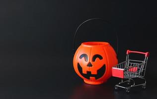 plastic Halloween pumpkin bucket  with shopping cart or trolley isolated  on black  background with copy space. Halloween holiday shopping  concept. photo