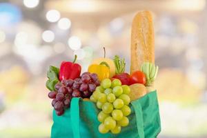 Fresh fruits and vegetables in reusable green shopping bag with supermarket grocery store blurred defocused background with bokeh light photo