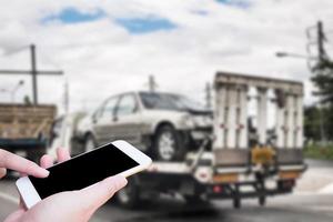 Hand using mobile smartphone for emergency roadside service with Broken car on tow truck after traffic accident background photo