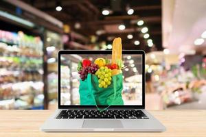 pasillo de supermercado fondo borroso con computadora portátil y bolsa de compras verde en mesa de madera concepto en línea de comestibles foto