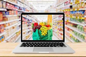 supermarket aisle blurred background with laptop computer and green shopping bag on wood table grocery online concept photo