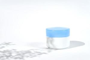 Cosmetic white jar blue lid with cream or lottion and flowers shadows. White background with daylight and harsh shadows photo