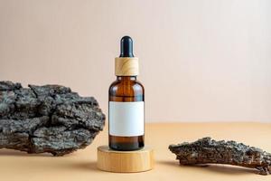 A mock-up of a brown cosmetics bottle with pipette and blank label on wooden podium with tree bark as decoration for cosmetics photography on beige background photo