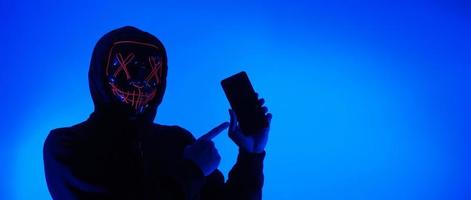 concepto de seguridad digital. hacker anónimo con máscara sosteniendo un teléfono inteligente pirateado. foto