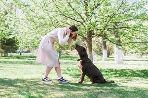 Woman and her puppy of chocolate Labrador retriever are walking in summer park. Owner and dog are looking into each others eyes. Friendship between human and dog. photo