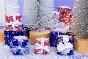 Gift boxes wrapped in shiny blue and red paper with artificial snow and Christmas trees on purple background. photo