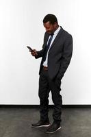 Smiling african american black man in business suit looks in smartphone white wall background photo