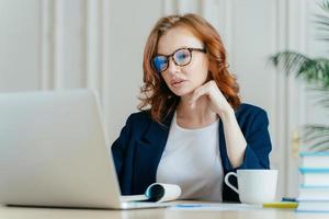 Shot of atttactive businesswoman concentrated in monitor of laptop computer, has serious focused gaze, wears optical glasses for vision correction, watches training video, drinks hot beverage photo