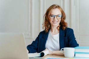 Glad smiling red haired young female concentrated on creating new business project, owns corporation, sits in front of laptop computer, wears optical glasses and elegant outfit, updates software photo
