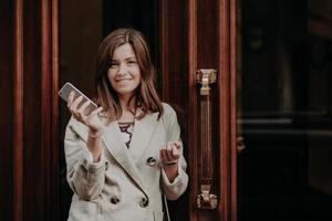 Elegant gorgeous lady in raincoat, holds mobile phone, waits for call, poses outdoor near doors, going for work, feels pleased, updates information on website, connected to high speed internet photo