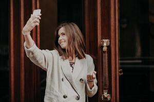 Pretty adorable woman makes selfie with smart phone, dressed in raincoat, poses outdoor, uses modern technology, shares photos in social networks. People, technology and entertainment concept