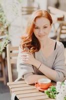 Vertical shot of attractive red haired young woman with wavy hair, dressed in warm jumper, drinks coffee or latte, sits at cafeteria outside, has pleasant conversation with boyfriend or lover. photo