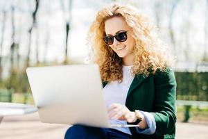 Young woman with sunglasses having fluffy hair smiling joyfully resting in park browsing internet using laptop computer surfing in social networks. People, lifestyle, technology and rest concept photo