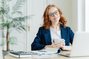 una maestra complacida hace trabajos de investigación en una computadora portátil, escribe información en el bloc de notas, disfruta tomando café, tiene una sonrisa feliz en la cara, se sienta en una habitación espaciosa en la planta de escritorio en segundo plano foto