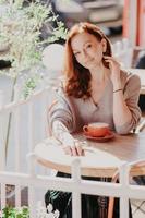 Photo of good looking ginger young woman in spectacles watches tutorial video online, poses in comfortable couch in modern apartment, dressed in elegant outfit, enjoys free internet connection