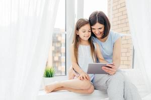 personas, tecnología, familia, concepto de niños. otro joven positivo y su pequeña hija se sientan en el alféizar de la ventana, sostienen una tableta digital, miran dibujos animados interesantes, posan cerca de la ventana con cortinas blancas foto