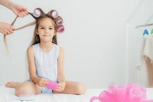 una niña atractiva tiene rulos en la cabeza, va a tener un peinado maravilloso, recibe lecciones de belleza de una madre cariñosa, aislada sobre fondo blanco. niños y concepto de belleza. foto