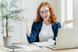 Image of cheerful redhead businesswoman watches webinar or tutorial video, uses free internet connection, raises palms with hesitation, sits at desktop with papers. Female owner of small business photo