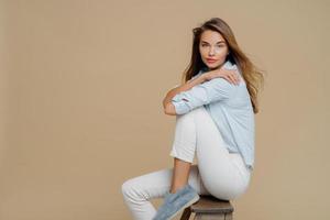 foto de estudio de una mujer caucásica hermosa y tranquila sentada en una silla, usa camisa, pantalones blancos y zapatos, mantiene las manos cruzadas sobre el cuerpo, tiene una mirada segura a la cámara, aislada sobre un fondo marrón.