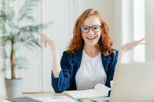 Shot of positive red haired female freelancer works remotely with paper documents, spreads hands to show her good result, sits at desktop with notepad and laptop computer, prepares course work photo