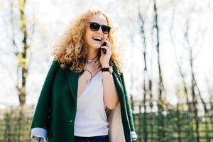 retrato de medio perfil de mujer atractiva con cabello rizado usando ropa elegante y gafas de sol hablando por teléfono inteligente, sonriendo y riendo con expresión feliz y emocionada tocando su cuello foto