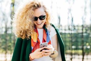 Beautiful female in fashionable round shades texting messages to her friends via social networks while standing outside in green park enjoying good sunny weather. People, communication, lifestyle photo