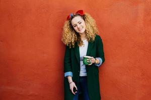 Young fashionable Caucasian woman with curly hair wearing headband and stylish jacket holding green cup of coffee having happy expression while resting against orange background. Woman with coffee mug