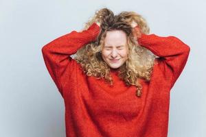 Stressful frustrated blonde woman tears out hair, regrets wrong doing, expresses negative emotions, wears loose red sweater, isolated over white studio background. Emotional young female indoor photo