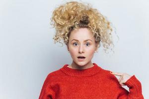 Shocked emotional amazed female stares at camera, indicates at sweater for your advertisment or information, isolated over white background. Surprised curly young woman isolated on white wall photo
