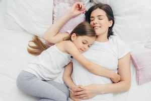 Top view of cheerful mother sleeps on white bed near her daughter who embraces mum with great love, shows good attitude, have pleasant dreams and nice rest, wears pyjamas. People, sleeping concept photo