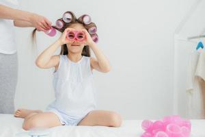 la foto horizontal de un hermoso niño pequeño tiene rulos en la cabeza, juega con ellos, tendrá un peinado maravilloso en el carnaval, aislado sobre fondo blanco. hija divertida juega junto con mamá