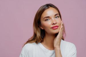 Headshot of lovely female model touches gently cheek, enjoys delicate face skin, wears makeup, has manicure, cares about her beauty, looks straightly at camera, dressed in white jumper, models indoor photo